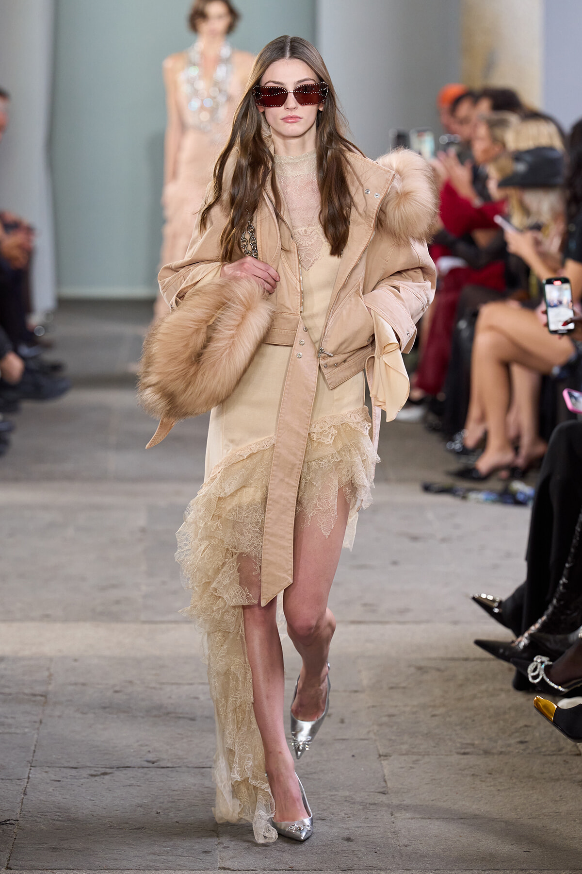 Model on a runway wearing a beige fur-trimmed coat over a lace dress, silver heels, and oversized sunglasses, carrying a large fur stole.