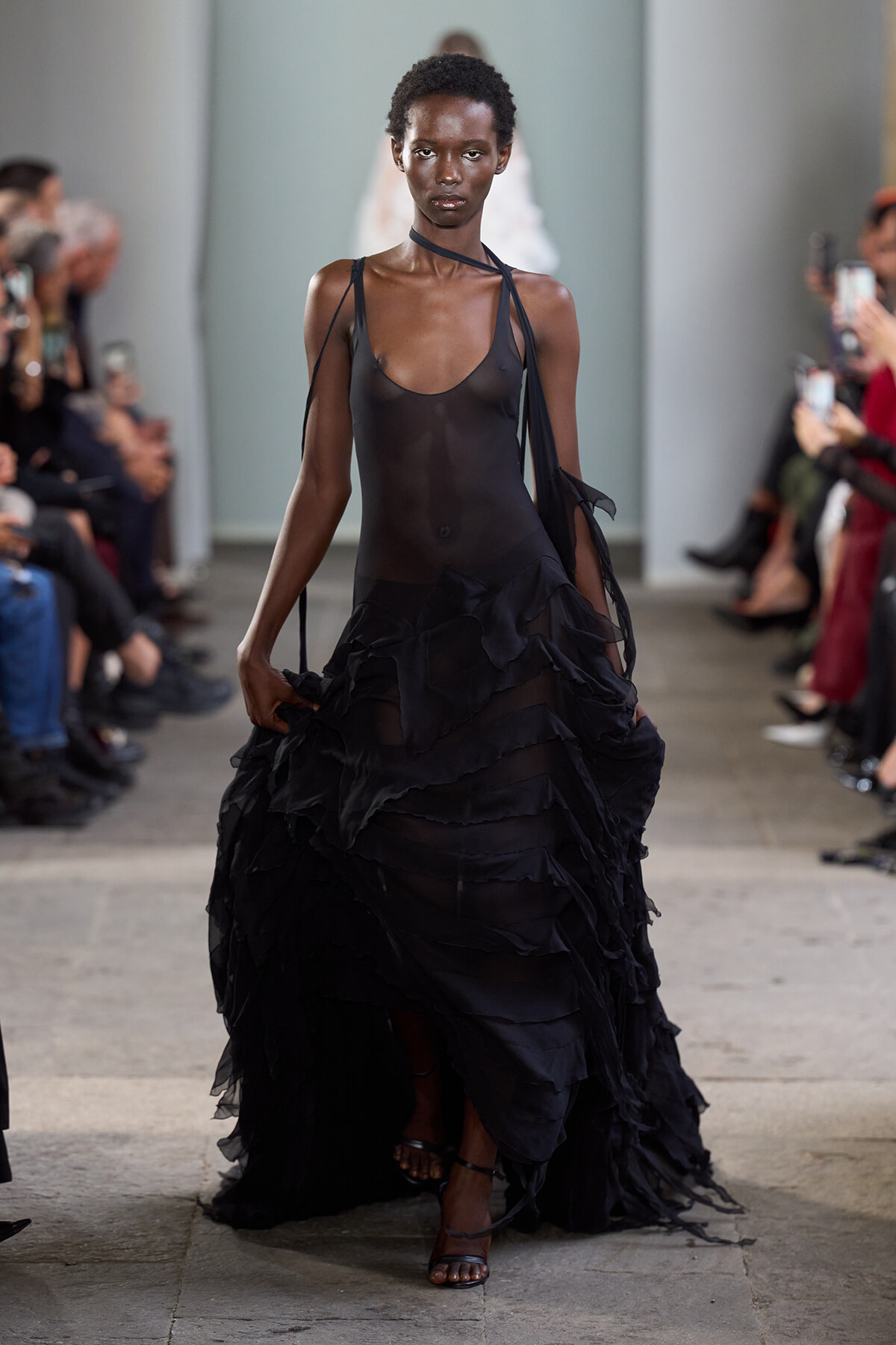 Model walking a runway in a sheer black halter dress with a layered, ruffled skirt, flanked by seated spectators on both sides.