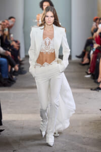 Model on a runway wearing a white tailored suit with a lace bralette and high-waisted pants; long white coat flowing behind.