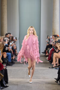 Model in a pink, layered ruffled dress walks down a fashion-show runway as audience members on both sides film with their phones.