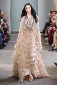 Model walking the runway in a cream tiered ruffle gown with long silver chain necklaces and cross pendants.