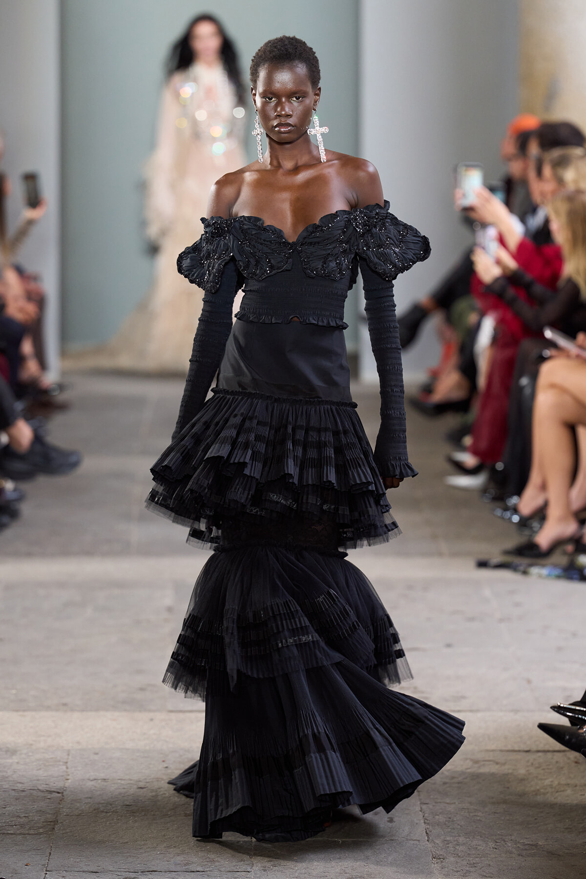 Model walks the runway in a black off-the-shoulder, ruffled gown with long sleeves and large earrings.