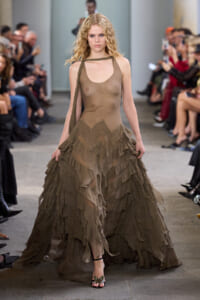 Model walking a runway in a sheer taupe dress with layered, ruffled skirt and strappy sandals.