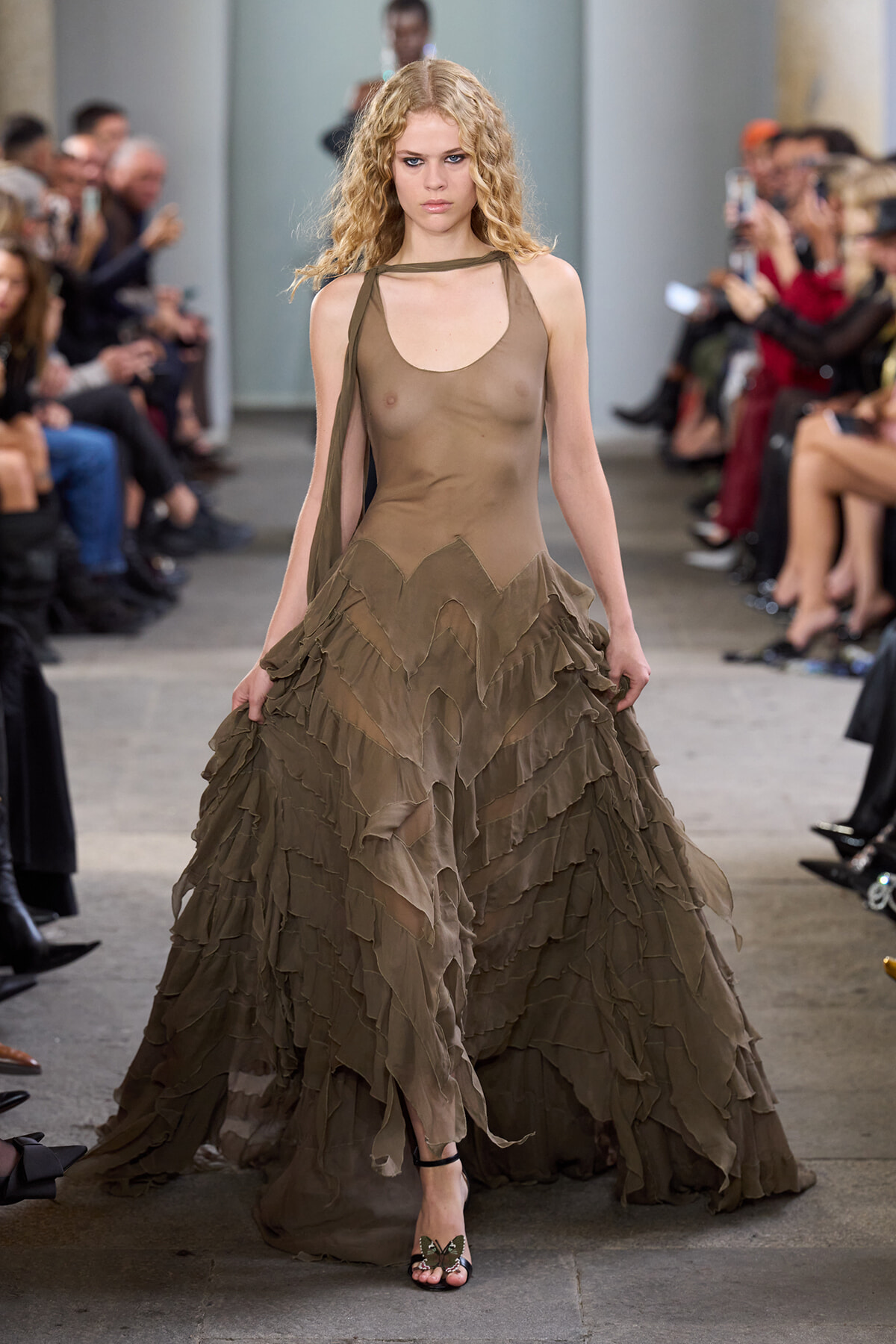 Model walking a runway in a sheer taupe dress with layered, ruffled skirt and strappy sandals.