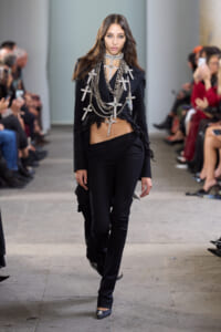 Model walking a fashion runway in a black tailored suit with multiple silver chain cross necklaces draped over the chest and shoulders.