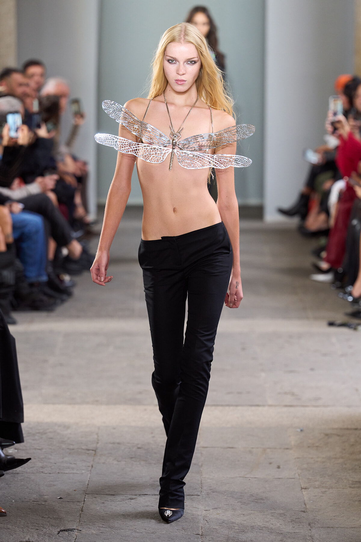 Runway model wearing a large metallic dragonfly-wing accessory across the chest, paired with black pants, walking the catwalk.