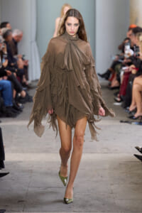 Model walks a fashion show wearing a taupe layered chiffon dress with a bow at the neck.
