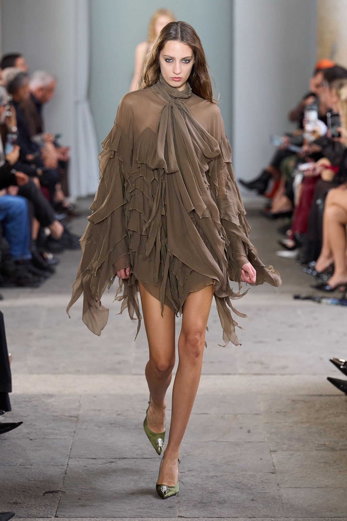 Model walks a fashion show wearing a taupe layered chiffon dress with a bow at the neck.