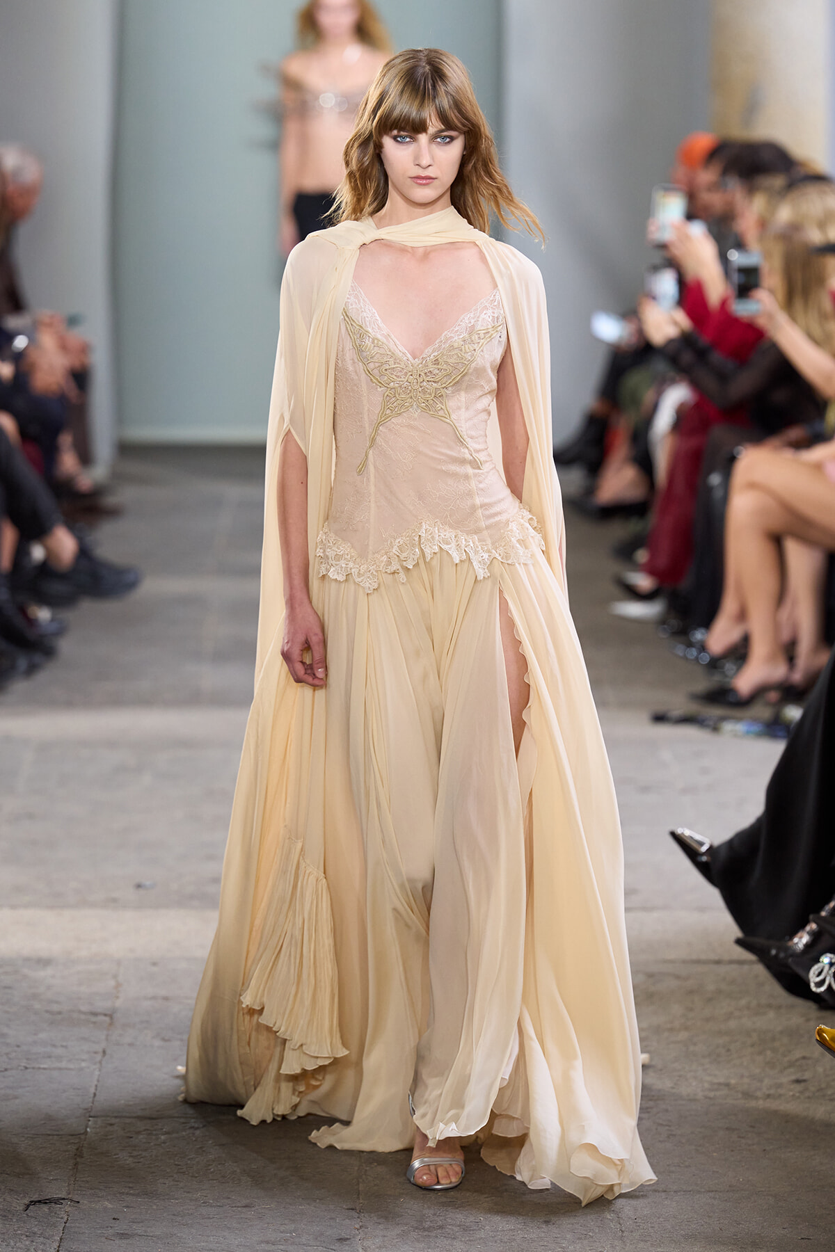 Model on a runway wearing a pale cream lace gown with a sheer cape, walking the catwalk.