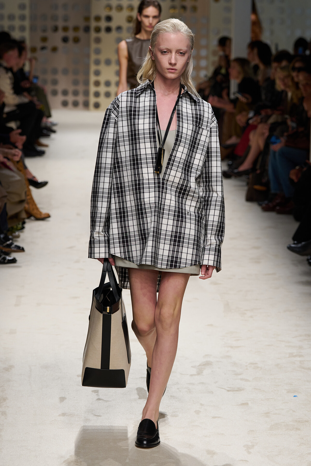 Model walks the runway in a black-and-white plaid oversized blazer over a beige dress, carrying a beige and black tote bag and wearing black loafers.