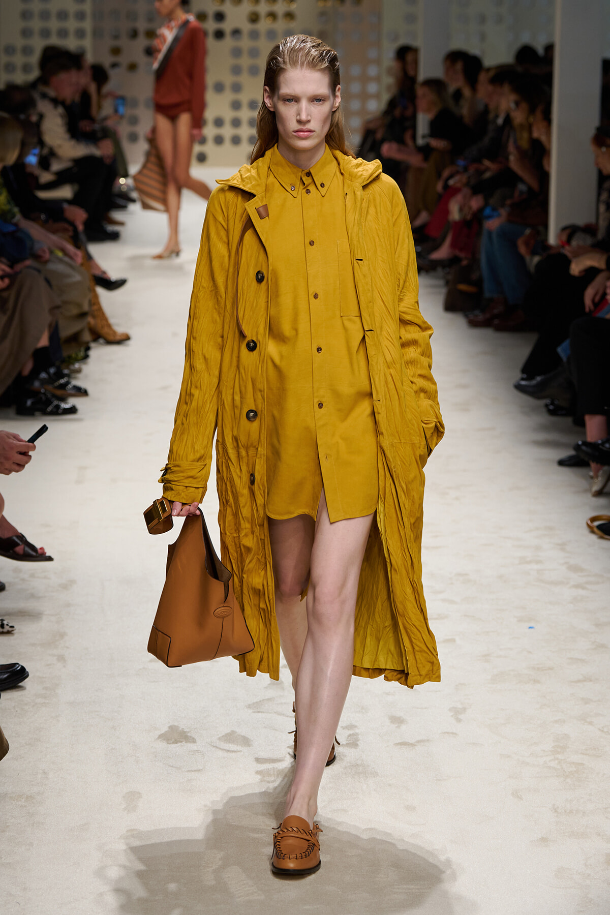 Model walks the runway in a mustard yellow trench coat over a matching shirt dress, carrying a brown tote, with seated audience in the background.