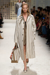 Model walking a runway in a long beige-and-white striped trench coat carrying a matching striped tote bag.
