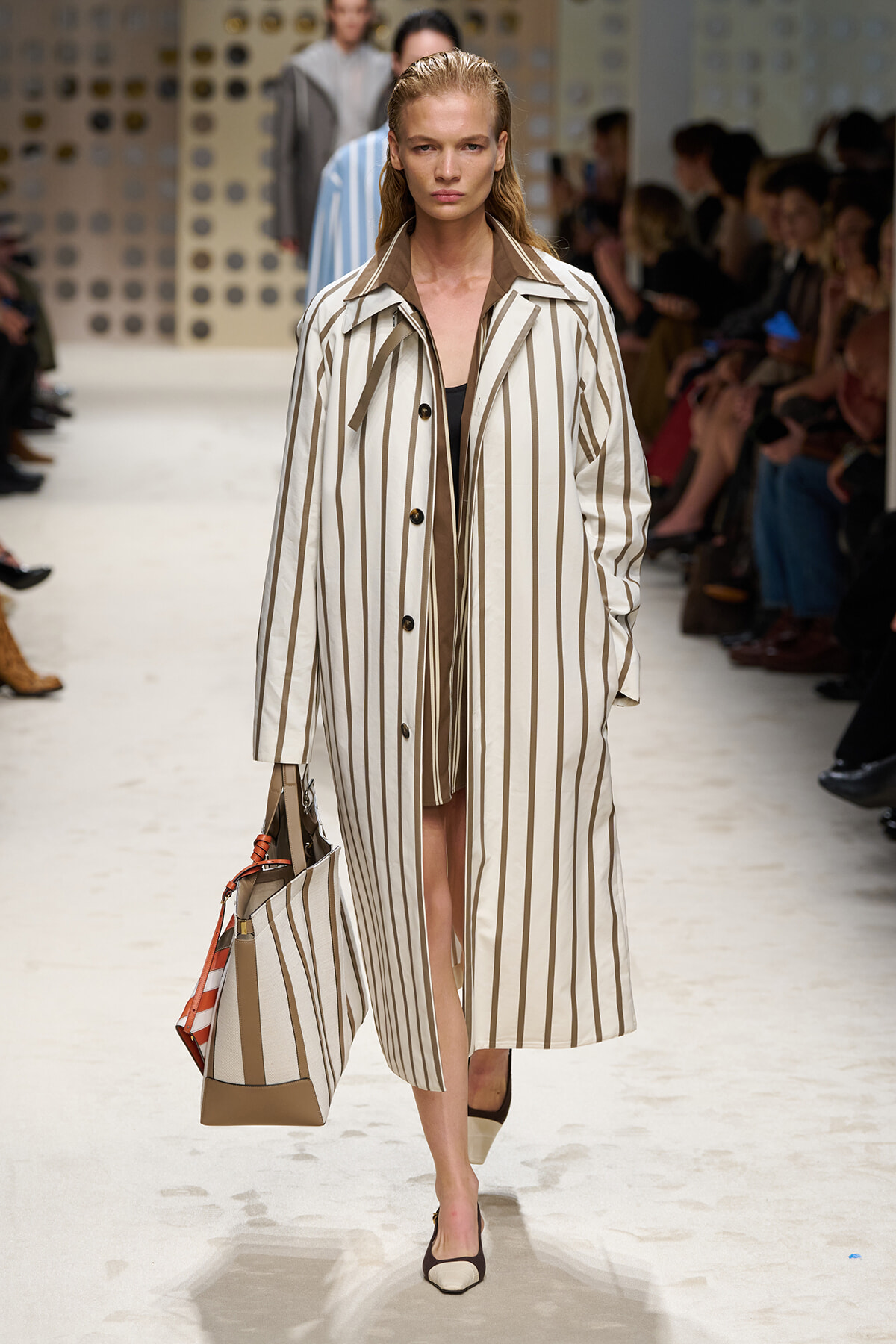 Model walking a runway in a long beige-and-white striped trench coat carrying a matching striped tote bag.
