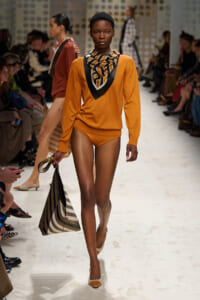 Model walking a fashion show runway in an orange top and matching bottoms with a striped scarf, carrying a striped tote bag.