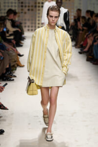 Model walks the runway in a pale yellow shift dress under a yellow striped button-down shirt, carrying a small yellow handbag.