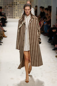 Model walks the runway wearing a long brown coat with bold white vertical stripes over a white shirt dress; hands in pockets, confident expression.