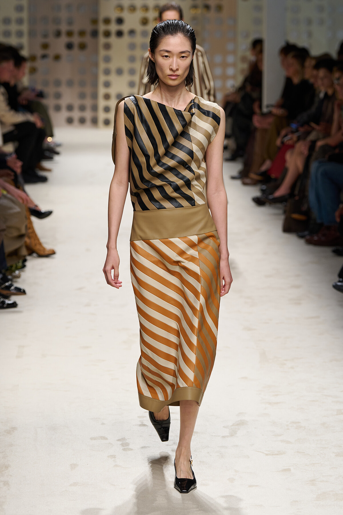 Model walks a runway in a color-block striped dress: diagonal black and gold top with a beige waist, and a tan-and-cream diagonal skirt with black heels on a white catwalk.