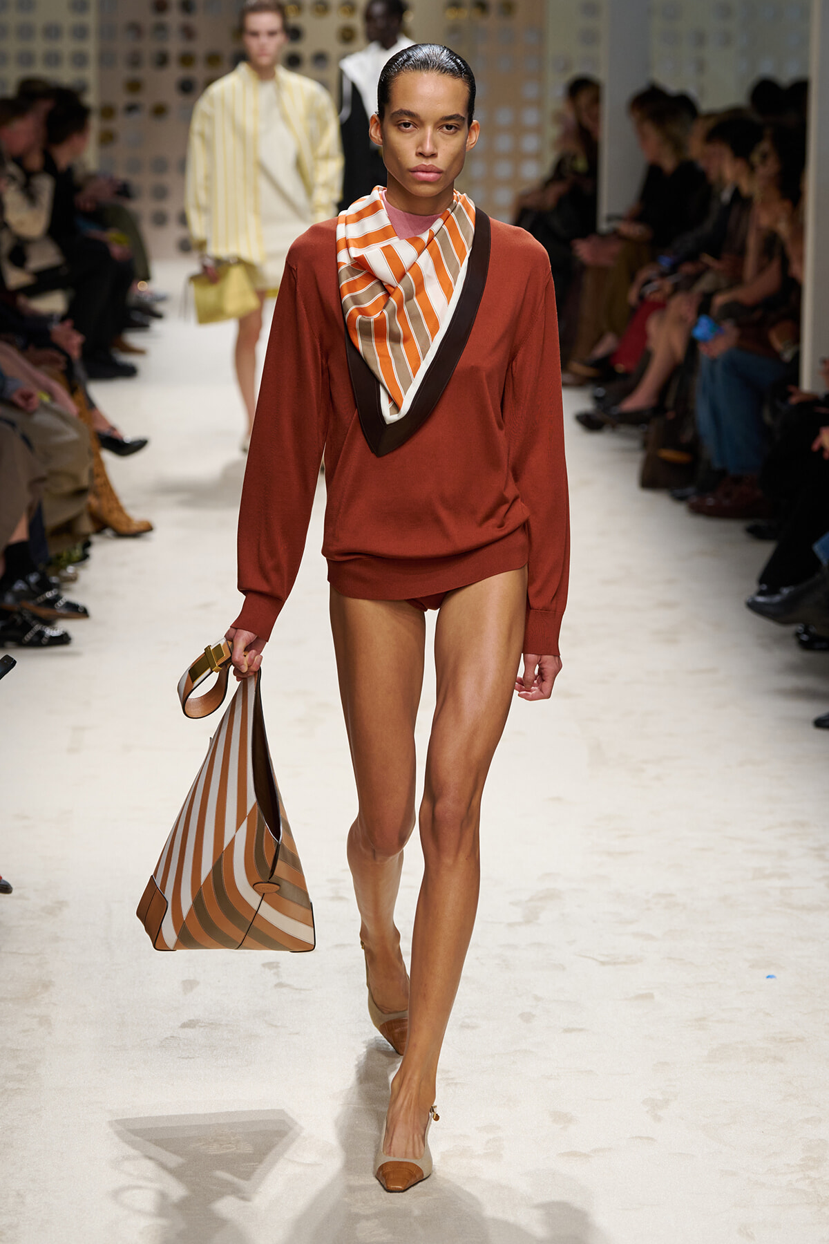 Model walking a fashion runway in a rust sweater with a striped orange-and-cream scarf, carrying a matching striped tote bag in brown tones.