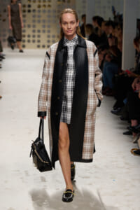 Model walking a fashion runway in a beige plaid coat with black leather panels, carrying a black handbag.