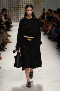 Model walks the runway in an all-black textured coat and dress, with a gold belt buckle, black bag, and black loafers with gold accents.