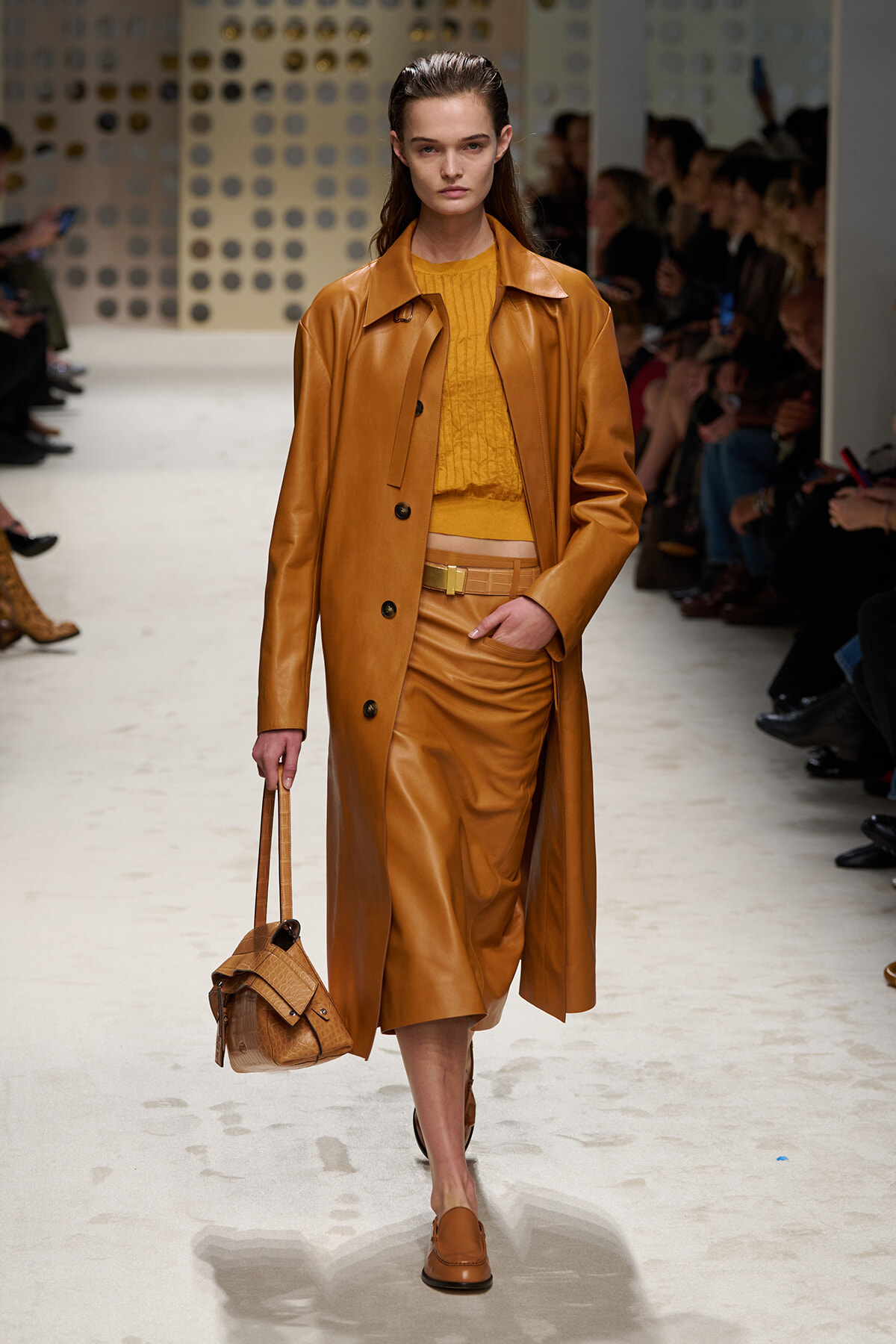 Model walks the runway in a caramel leather coat over a mustard knit top and skirt, carrying a tan handbag.