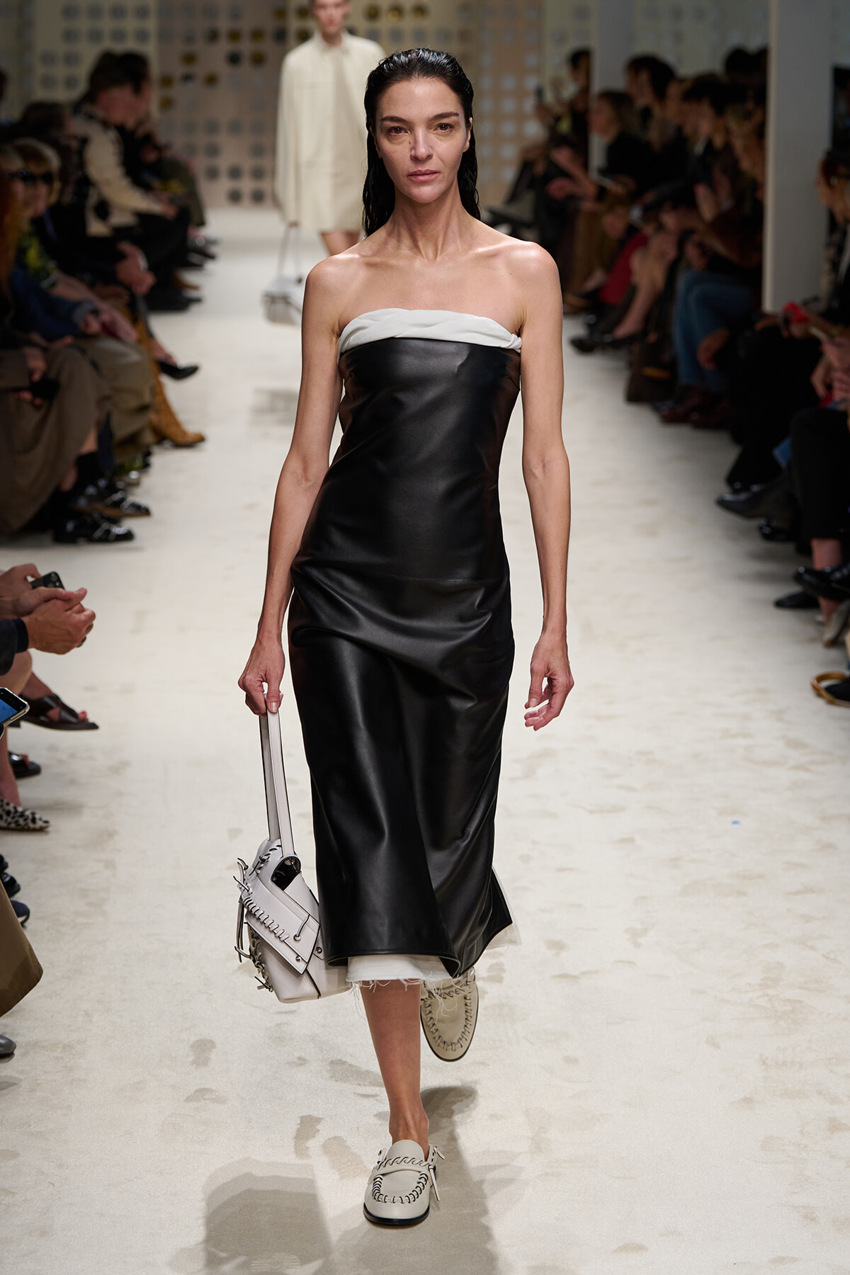 Model walking a runway in a strapless black satin midi dress with a white top, carrying a light-gray handbag.