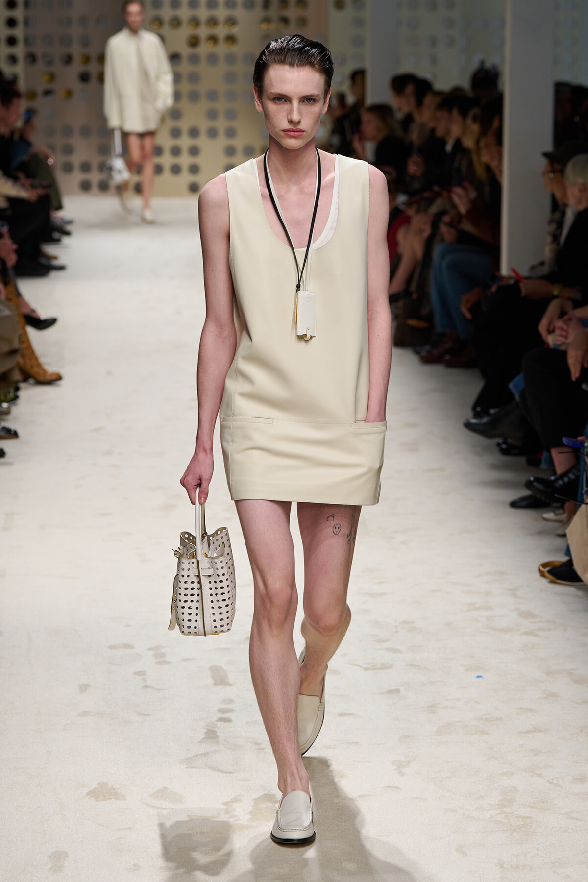 Model walks the runway in a sleeveless cream mini dress with front pockets, carrying a perforated beige handbag and wearing cream loafers.