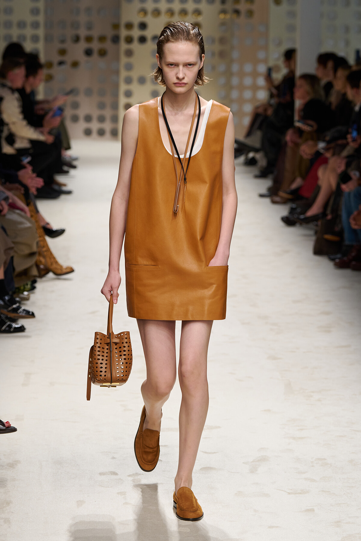 Model on a runway wearing a caramel leather shift dress with matching loafers and a perforated tan tote bag.