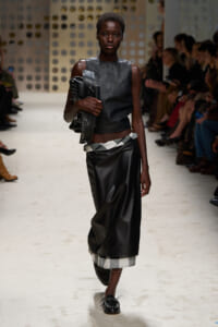 Runway model in a sleeveless black leather top and long black leather skirt with a black-and-white checkered hem, holding a large black bag under her arm as spectators watch.