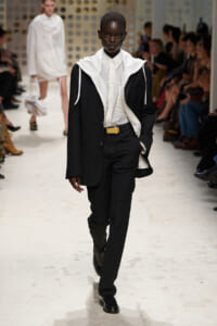Model walks a fashion runway in a black suit with a white oversized collared inner layer and hooded details, audience seated along the sides.