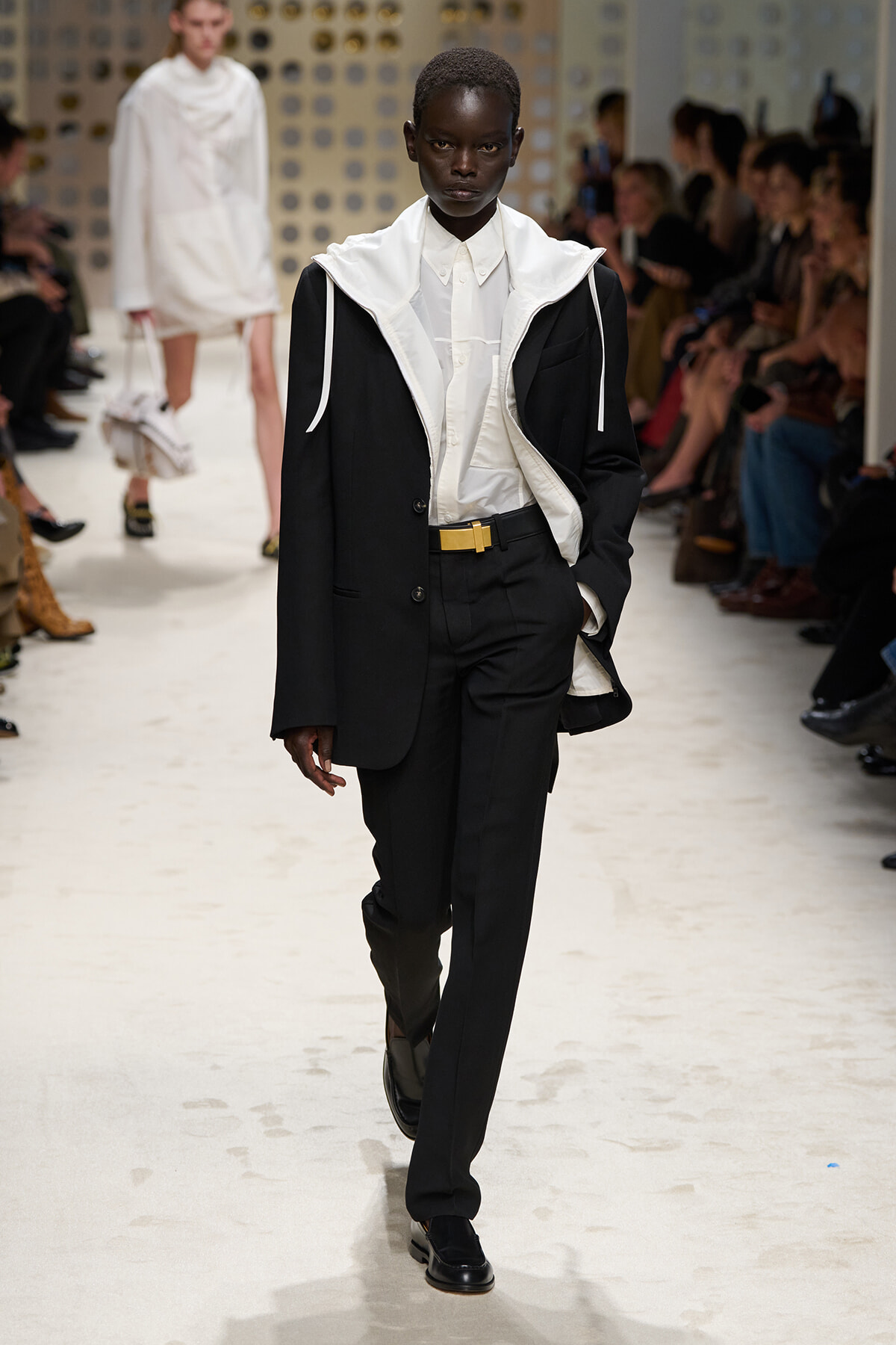 Model walks a fashion runway in a black suit with a white oversized collared inner layer and hooded details, audience seated along the sides.