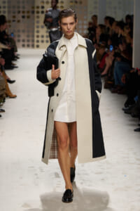 Model walking a fashion runway in a cream-and-black color-block trench over a white shirt dress, holding a black clutch.
