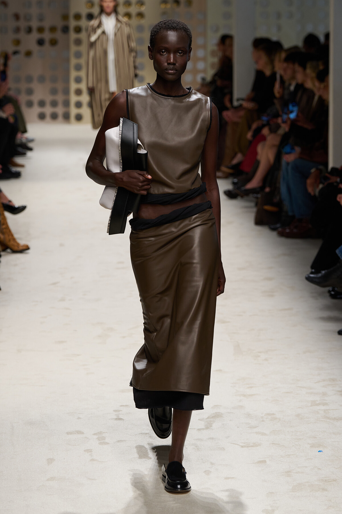 Female model walks the runway in a metallic brown leather sleeveless top and matching long skirt, with a black wrapped belt and a stack of black and white clutch bags, as seated audience watch on both sides.