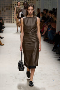 Model walks the runway in a brown leather sleeveless midi dress, carrying a black handbag; audience sits along the sides.