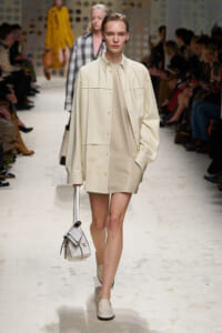 Female model walks the runway in a beige oversized shirt-jacket over a matching mini dress, carrying a light gray handbag.