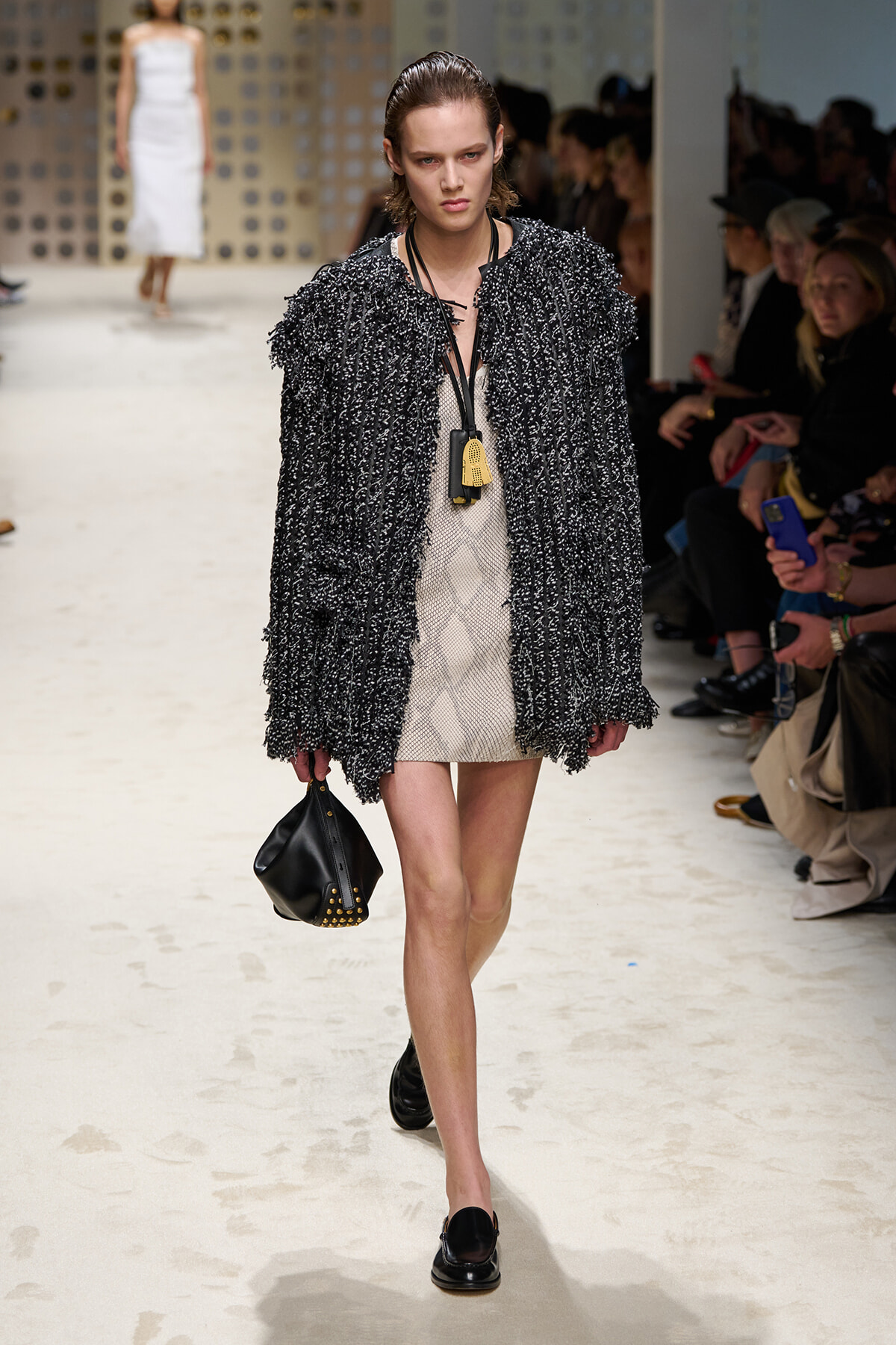 Model walking runway in a chunky black bouclé coat over a beige mini dress, carrying a studded black handbag.