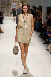 Female model walking the runway in a beige snakeskin-print sleeveless mini dress, white loafers, and a small taupe handbag; audience seated on sides.