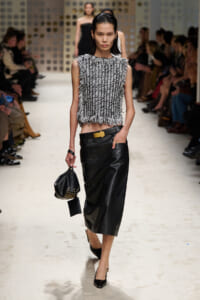 Model walks the runway wearing a textured sleeveless top and a black leather midi skirt, holding a studded black handbag.