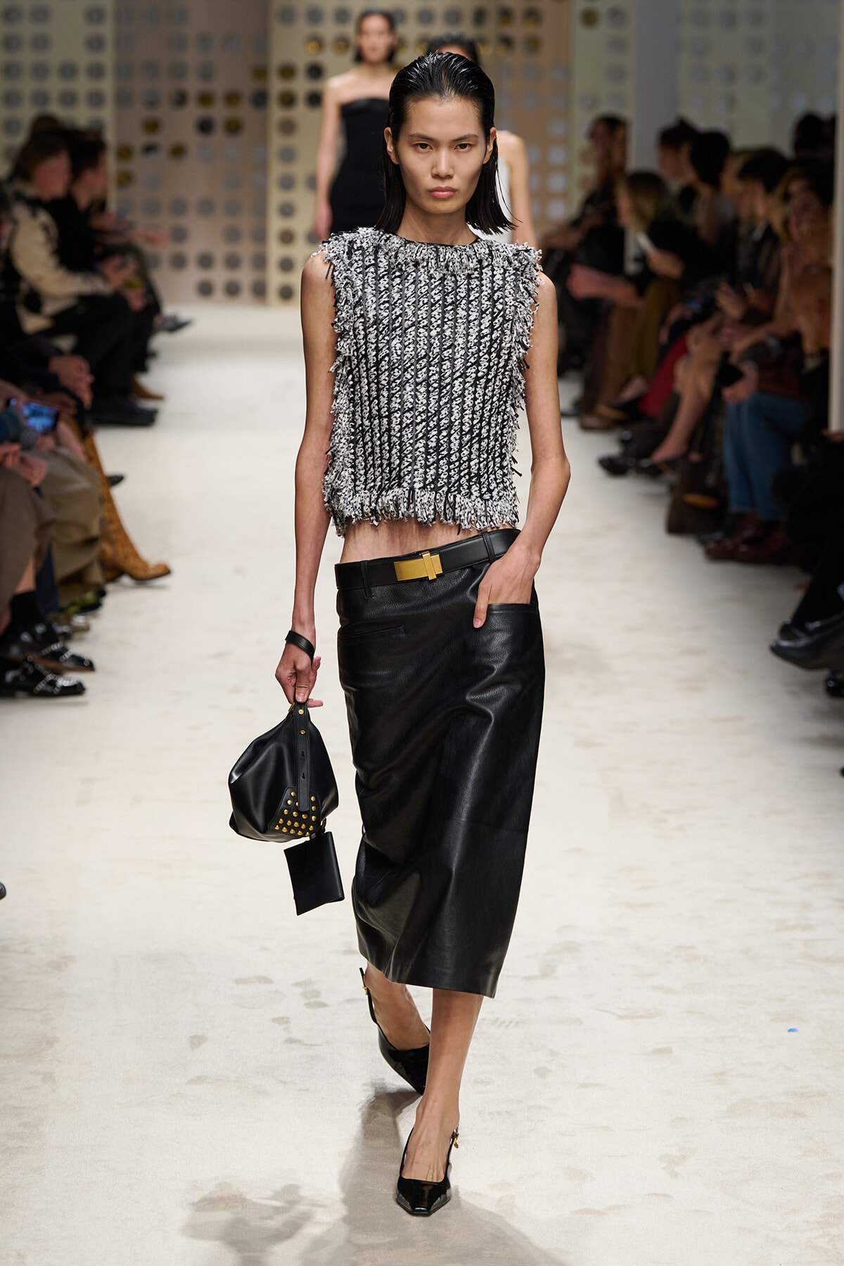 Model walks the runway wearing a textured sleeveless top and a black leather midi skirt, holding a studded black handbag.