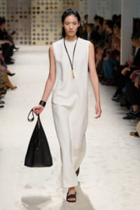 Model walking the runway in a sleeveless white textured jumpsuit, carrying a large black handbag and wearing a long pendant necklace.