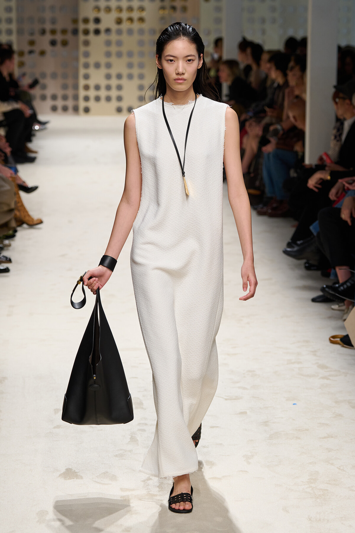 Model walking the runway in a sleeveless white textured jumpsuit, carrying a large black handbag and wearing a long pendant necklace.