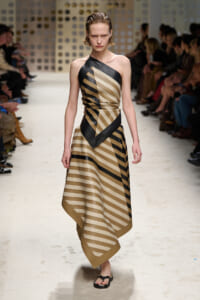 Model walking a fashion runway in a one-shoulder beige and black striped dress with angular black trim, paired with black sandals.