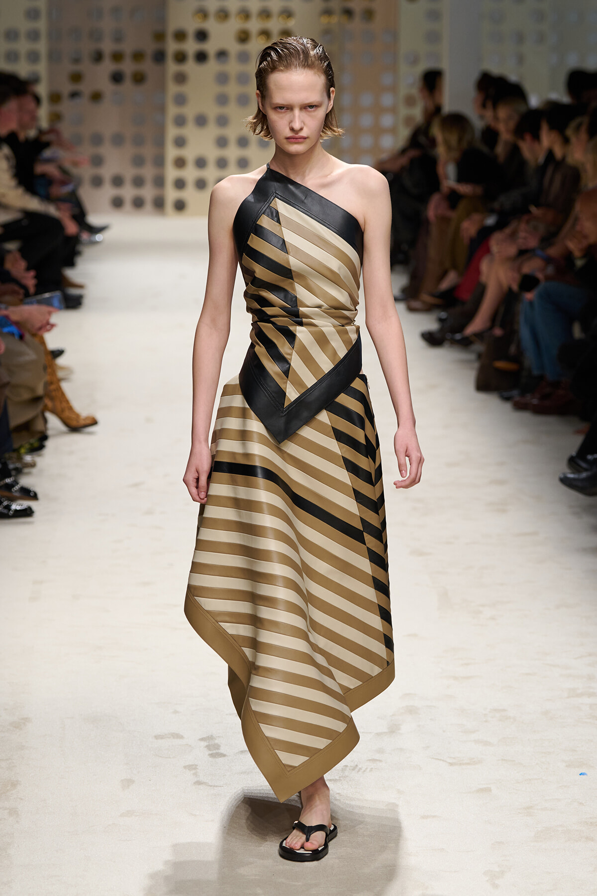 Model walking a fashion runway in a one-shoulder beige and black striped dress with angular black trim, paired with black sandals.