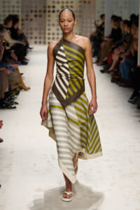 Model on a runway wearing a one-shoulder, olive-gray striped dress with asymmetric panels and white sandals.