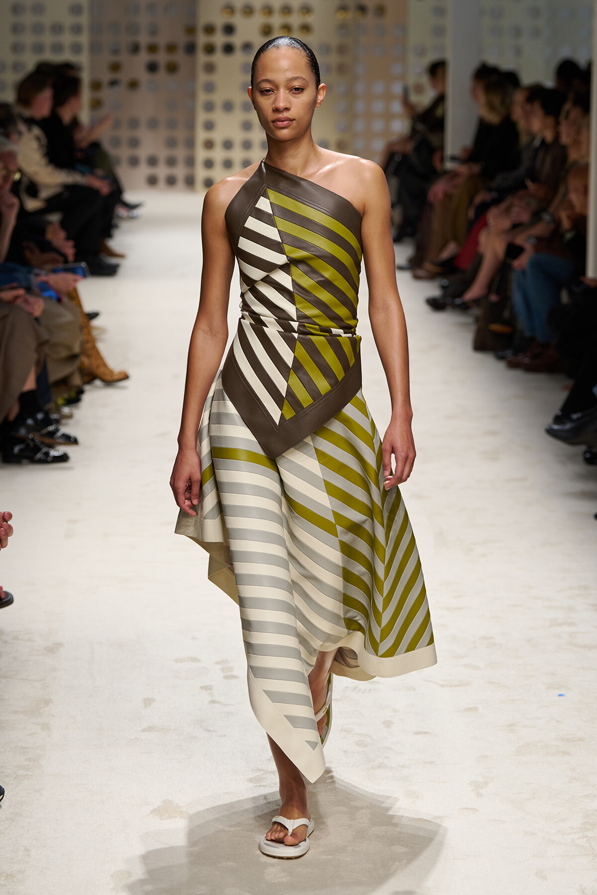 Model on a runway wearing a one-shoulder, olive-gray striped dress with asymmetric panels and white sandals.