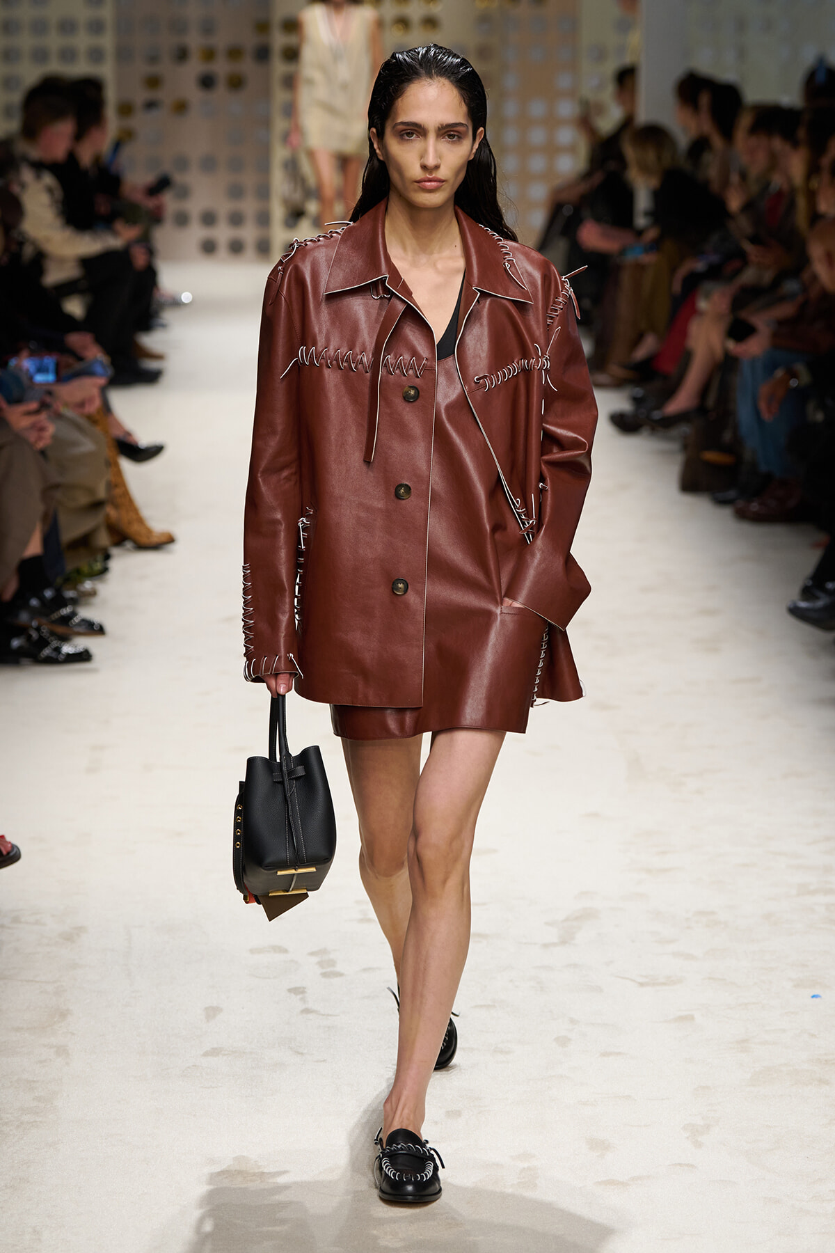 Model on a runway wearing a brown leather blazer with white stitch detailing and a matching mini skirt, carrying a black handbag.