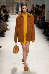 Model walks the runway in a warm-brown suede jacket over a mustard-yellow shirt and shorts, carrying a small tan handbag.