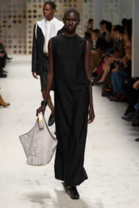 Model walks the runway in a sleeveless black maxi dress, carrying a large light-gray snakeskin tote with gold hardware.