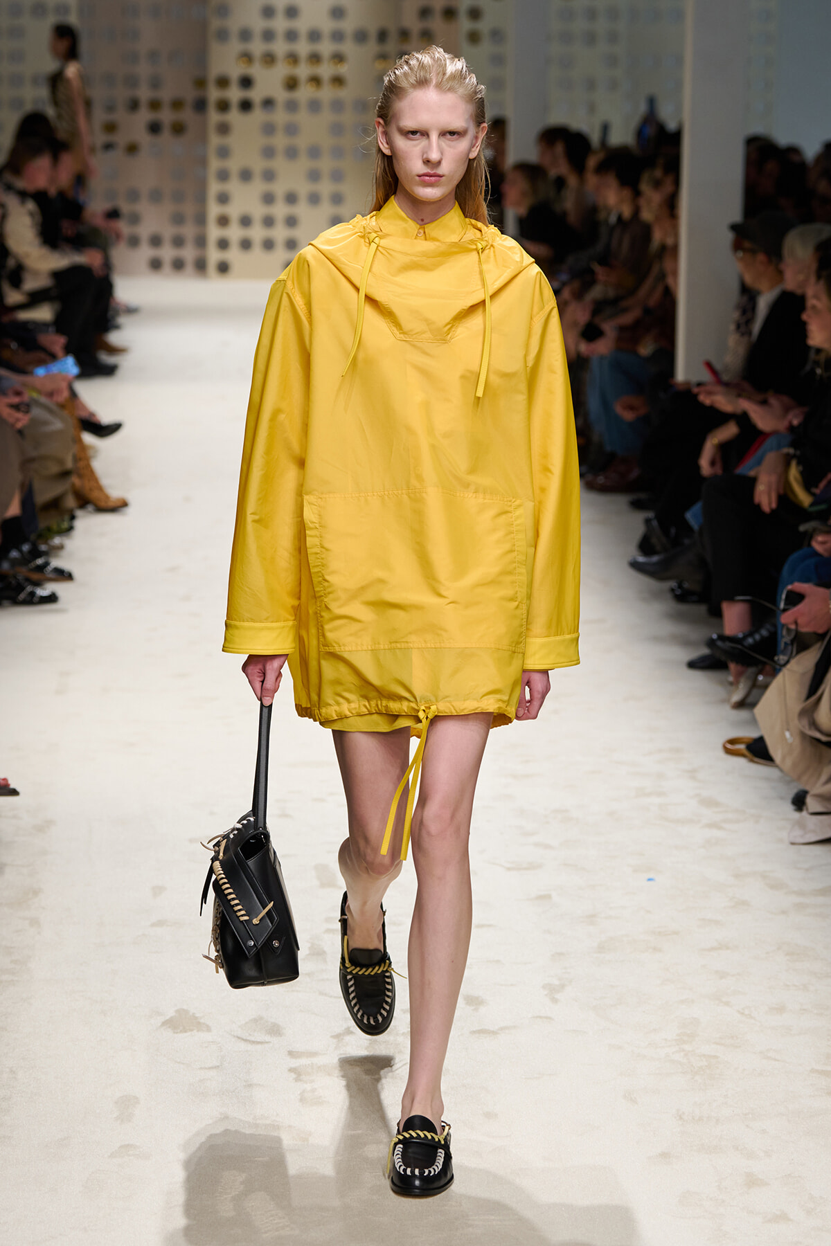 Model wearing an oversized yellow windbreaker with hood on a runway, carrying a black handbag.
