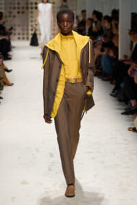 Model walking a fashion runway in a brown suit with a bright yellow inner jacket and collar, accessorized with a tan belt and loafers.